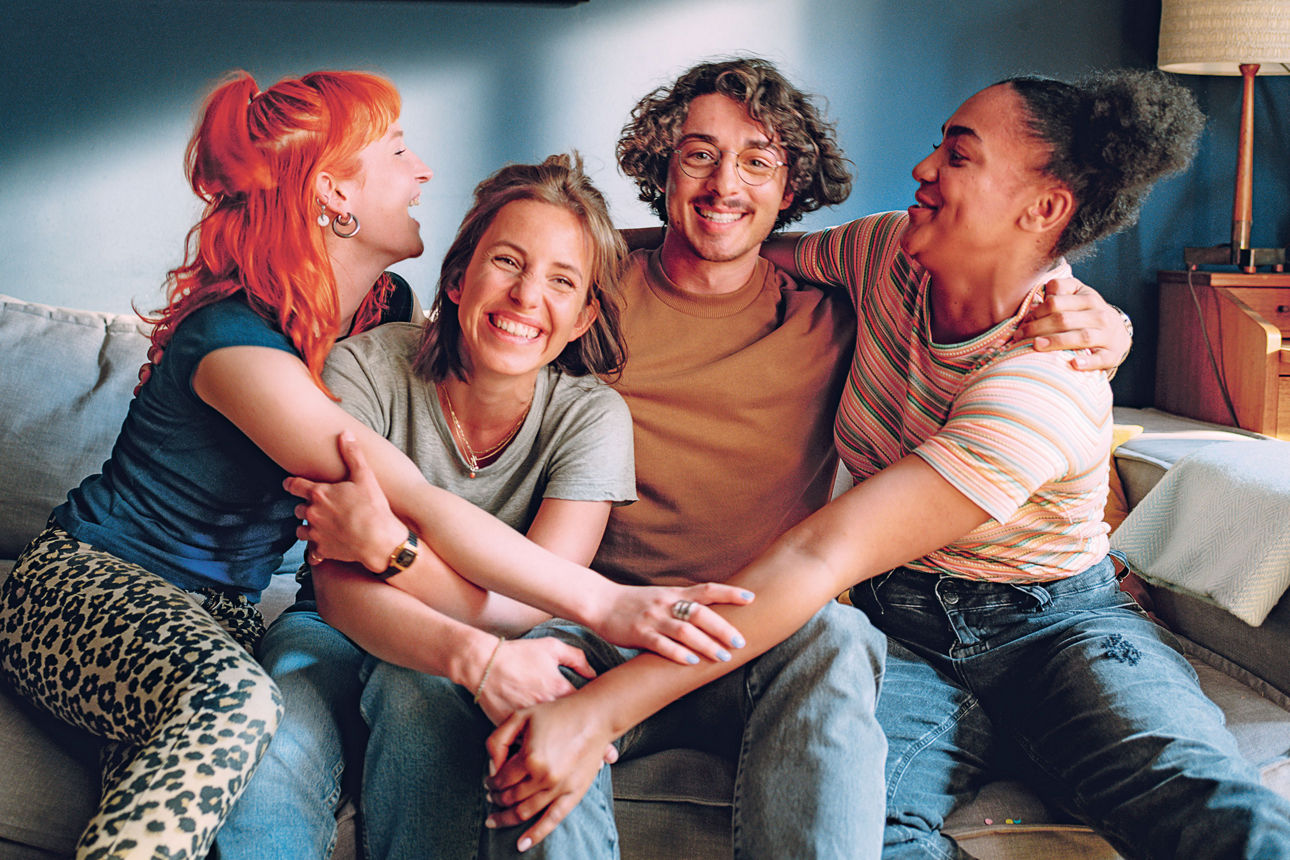 Vier junge Menschen sitzen auf einer Couch, nehmen sich in den Arm und lächeln.