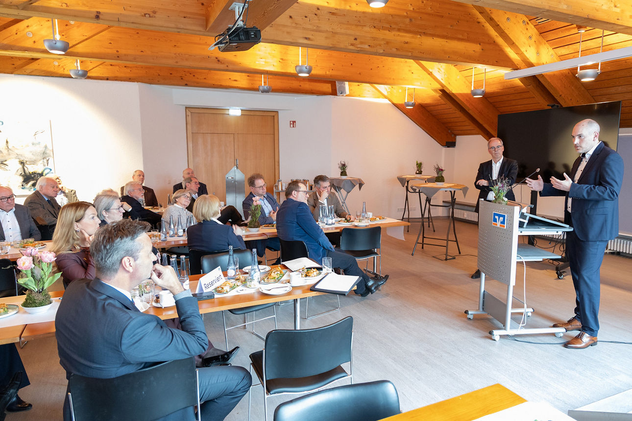 Der Polizeipräsident von Gelsenkirchen, Tim Frommeyer, steht an einem Rednerpult und spricht zu einer sitzenden Gruppe in einem Tagungsraum mit Holzdecke. Neben dem Redner steht unser Vorstand Ingo Abrahams und hört aufmerksam zu. Die Teilnehmenden sitzen an Tischen, auf denen Speisen sowie Unterlagen liegen. Die Atmosphäre wirkt angenehm locker und die Zuhörenden folgen dem Vortrag aufmerksam. 
