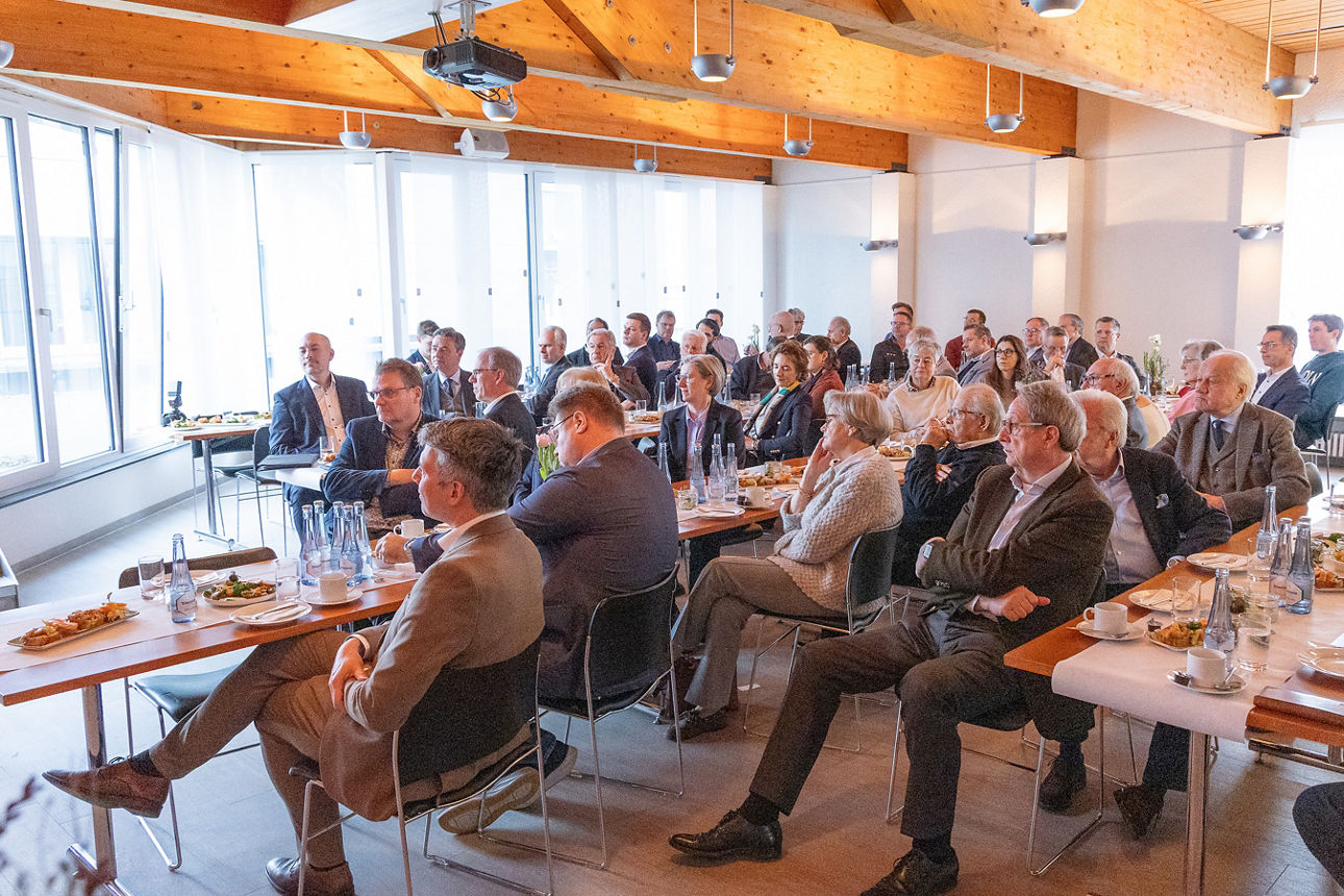 In einem hellen Konferenzraum sitzen zahlreiche Teilnehmende an langen Tischen und hören einer Präsentation oder Rede zu. Der Raum verfügt über große Fensterfronten, die viel Tageslicht hereinlassen, sowie eine Holzdecke mit sichtbaren Balken. Auf den Tischen stehen Getränke, Teller mit Speisen und Unterlagen. Die Anwesenden blicken geschlossen nach vorn in Richtung des Vortragenden oder der Präsentationsfläche.
