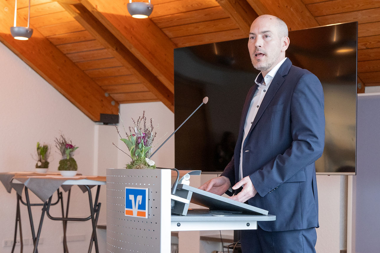 Der Polizeipräsident von Gelsenkirchen, Tim Frommeyer, steht an einem Rednerpult und spricht zu einer sitzenden Gruppe in einem Tagungsraum mit Holzdecke. Neben dem Redner steht unser Vorstand Ingo Abrahams und hört aufmerksam zu. Die Teilnehmenden sitzen an Tischen, auf denen Speisen sowie Unterlagen liegen. Die Atmosphäre wirkt angenehm locker und die Zuhörenden folgen dem Vortrag aufmerksam. 