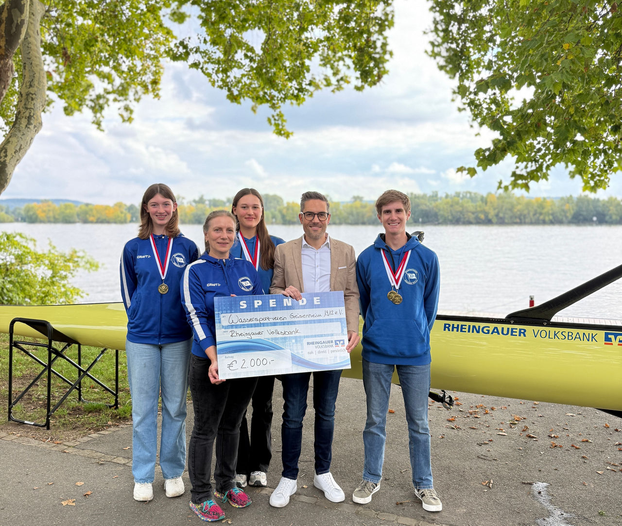 Gruppe von fünf Personen steht am Ufer vor einem gelben Ruderboot mit der Aufschrift ‚Rheingauer Volksbank‘, eine Person hält einen großen Spendenscheck über 2000 Euro.