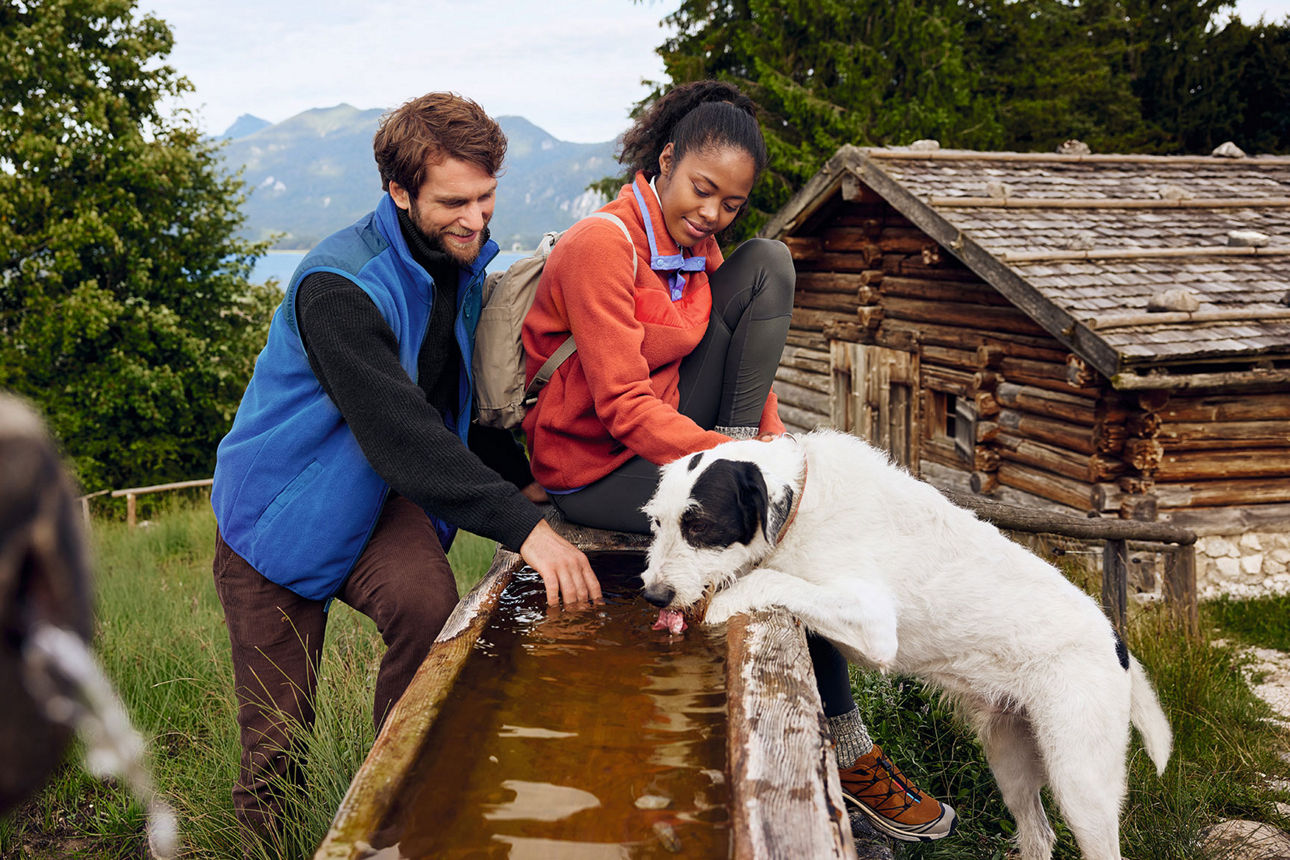 Ein Paar steht mit Hund an einem Brunnen in den Bergen.