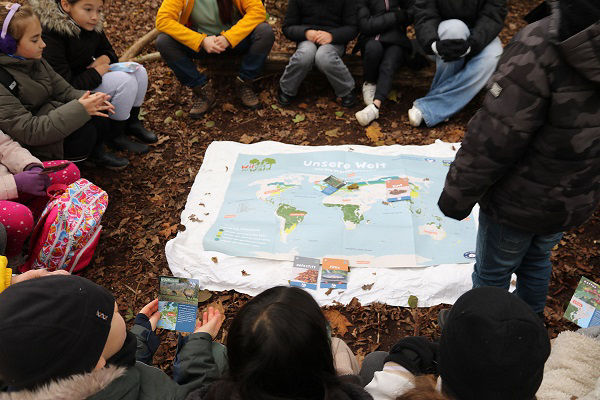 Kinder und Erwachsene sitzen im Kreis auf dem Boden. In der Mitte liegt eine große Weltkarte. Der Waldboden ist bedeckt mit Laub.
