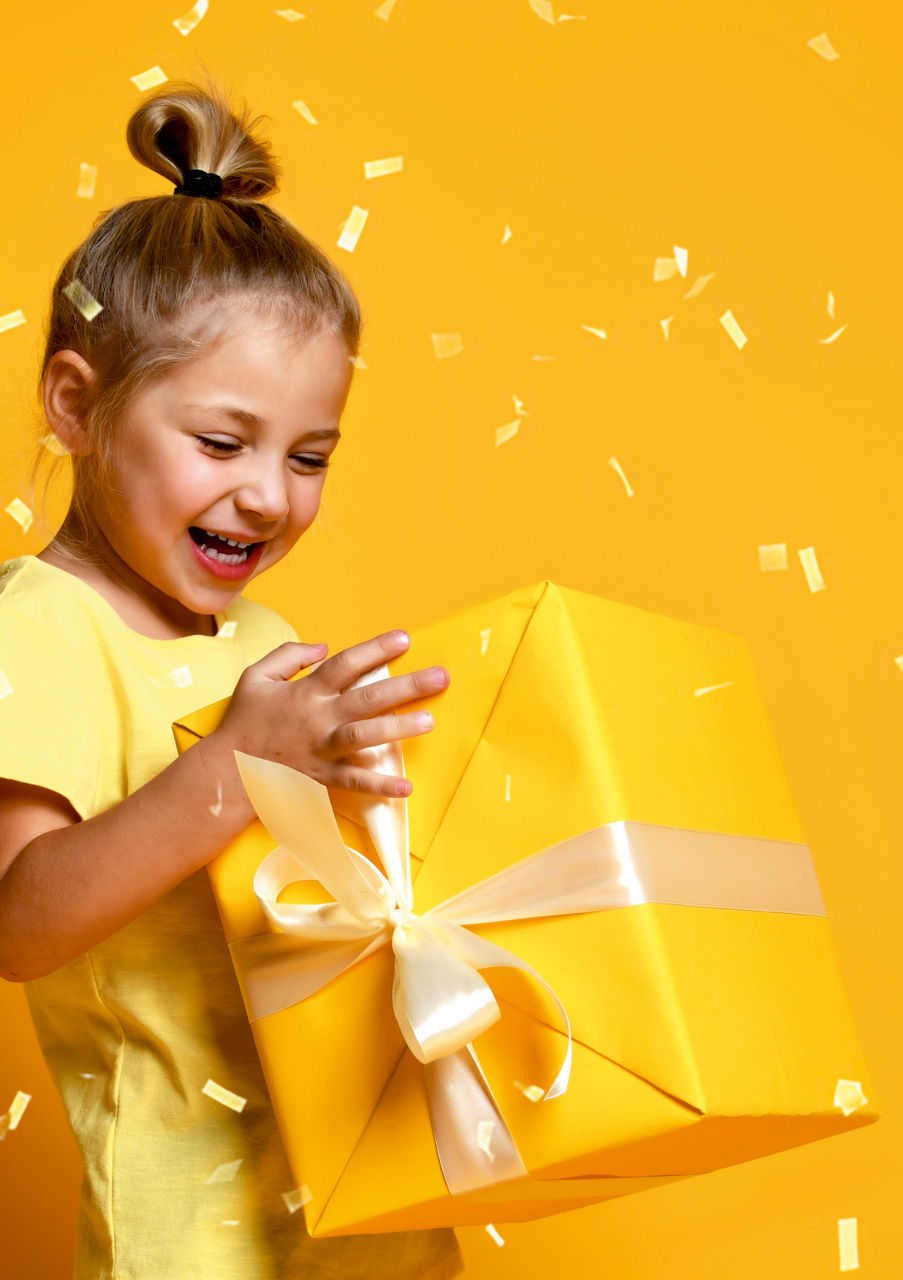 Ein kleines Mädchen hält ein Geschenk in der Hand, welches Gelb verpackt ist und Konfetti regent.