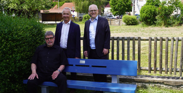 Drei Männer stehen vor einer blauen Bank in einem grasbewachsenen Park.
