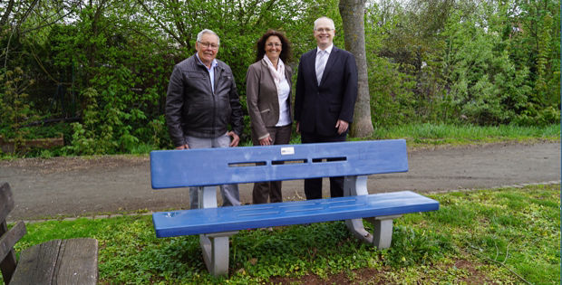 Drei Personen stehen vor einer blauen Bank in einem Park.