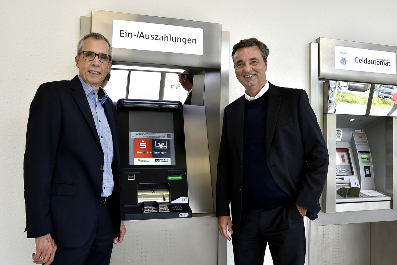Zwei Männer stehen vor einem Geldautomaten, auf dem das Sparkassen- und das Volksbank-Logo zu sehen ist.