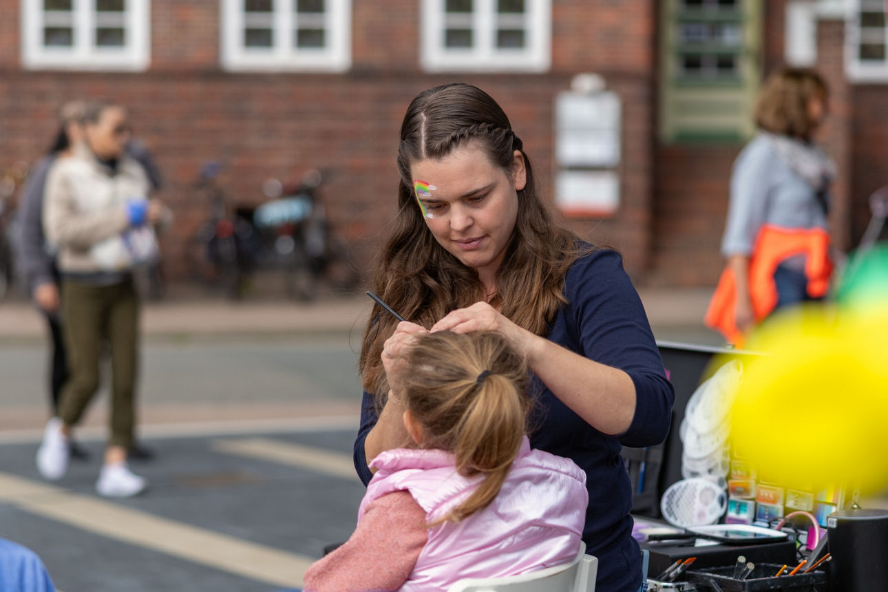 Eine junge Frau schminkt das Gesicht eines Kindes, im Vordergrund sind Luftballons zu sehen, im Hintergrund ein Haus
