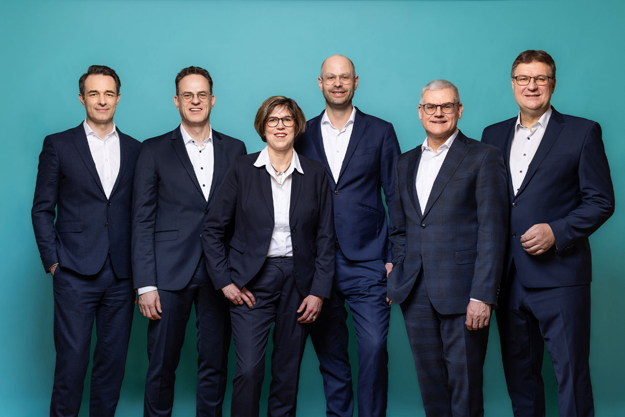 Der sechsköpfige Vorstand der VR RegionalBank nebeneinanderstehend. Der Hintergrund ist petrolfarben.