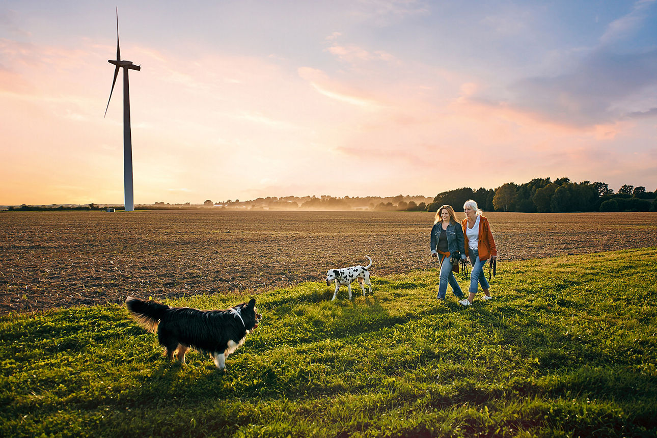 Zwei Frauen die mit einem Hund im Feld spazieren gehen. Im Hintergrund ist ein Windrad zu sehen.