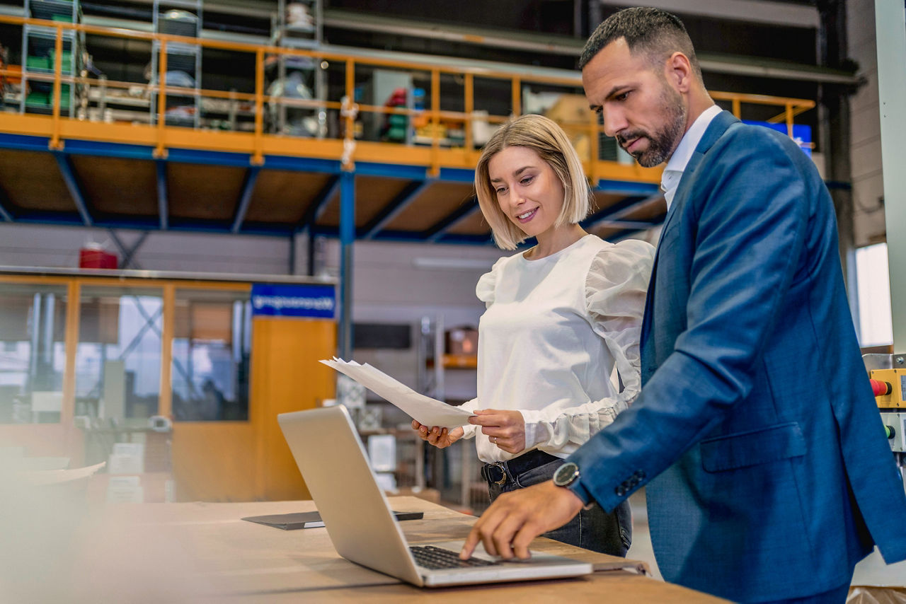 Eine Frau und ein Mann stehen vor einem Laptop in einer Fabrik