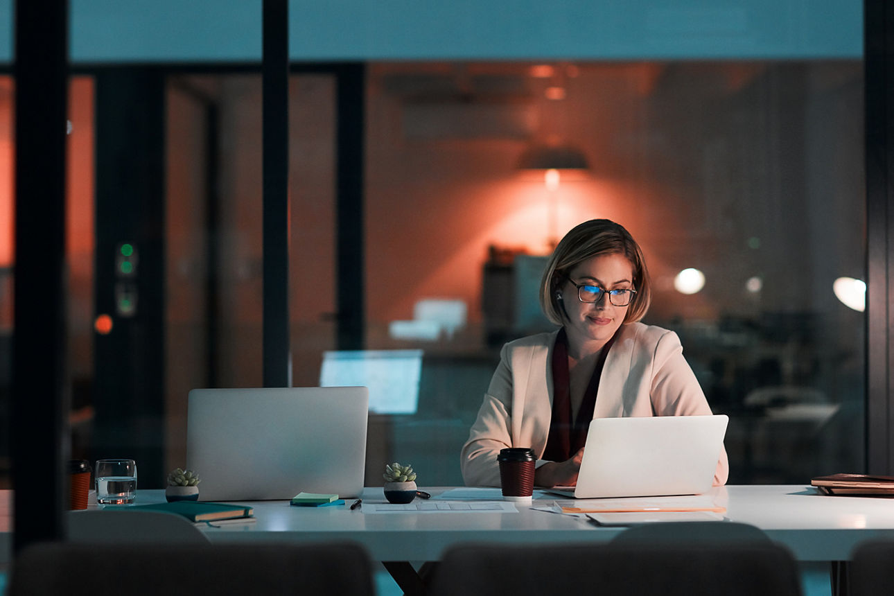 Eine Frau sitzt in einem etwas abgedunkelten Raum mit großer Fensterfront vor einem Laptop