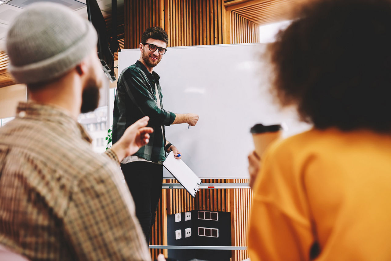Mann mit Brille vor Whiteboard und zwei Personen sitzend im Vordergrund