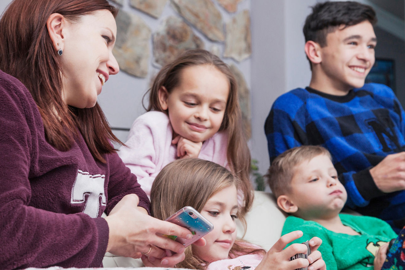 Eine Mutter und ihre 4 Kinder, zwei Mädchen und zwei Jungen im unterschiedlichen Alter, sitzen auf dem Sofa und schauen sich Sachen auf dem Handy an. Der älteste Sohn hat ein eigenes Handy auf das er schaut. Der kleine Bruder bläst gelangweilt Luft aus seinen Wangen. .