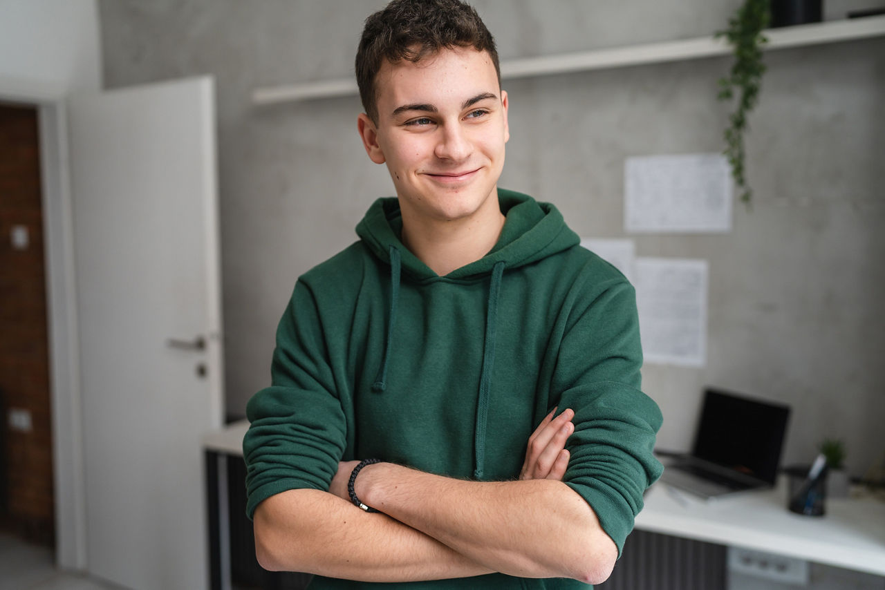 Ein junger Mann trägt einen grünen Pullover und steht vor seinem Schreibtisch