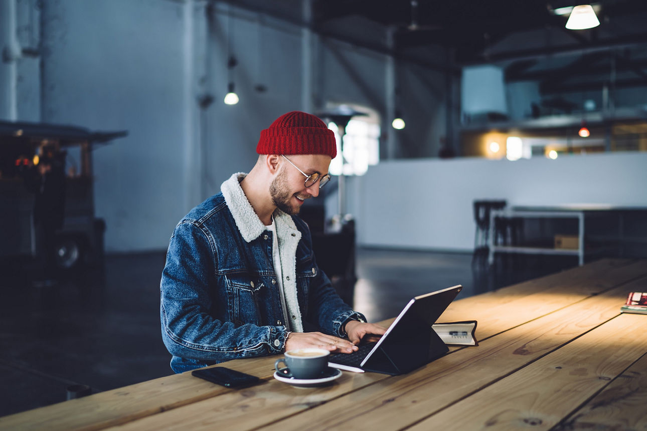 Person mit Laptop und Kaffe am langen Holztisch sitzend