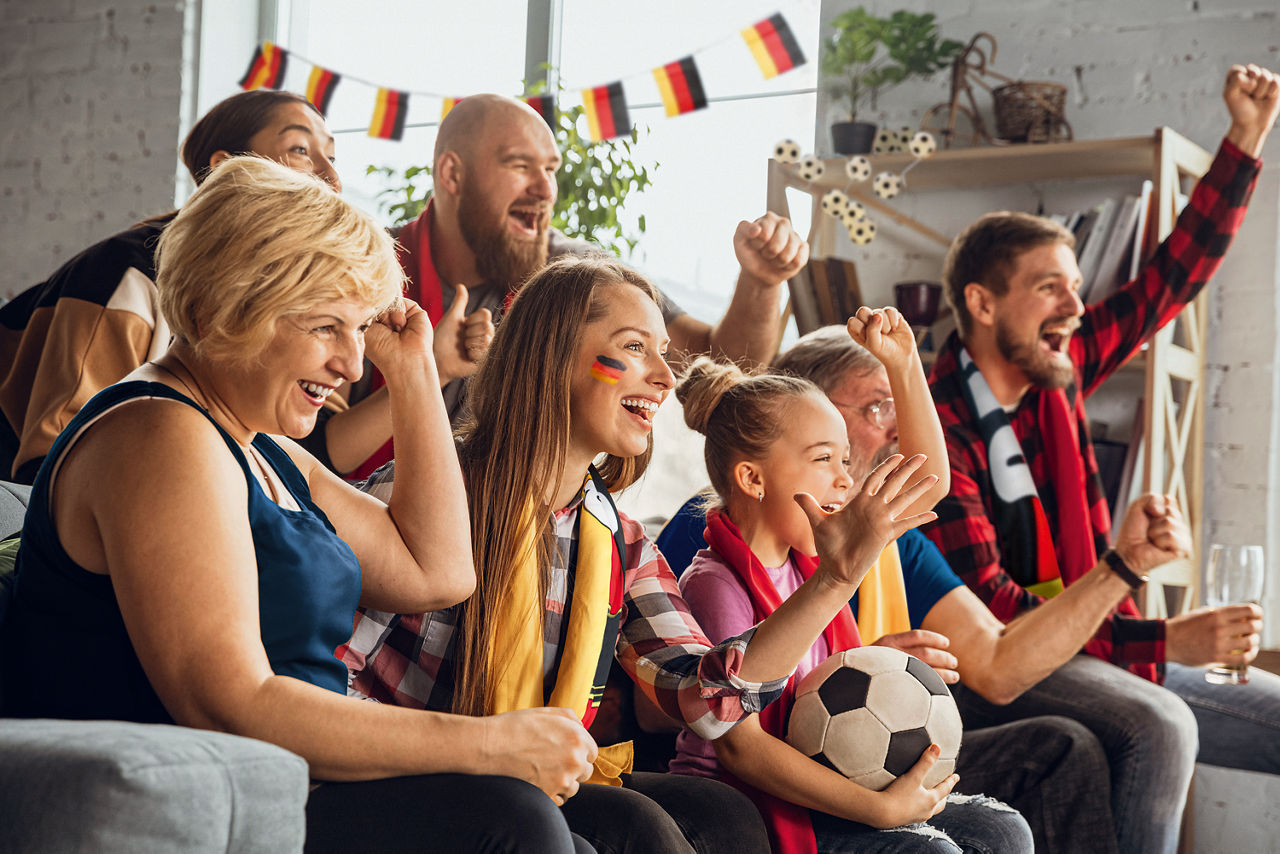 Menschen verschiedener Generationen sitzen in Fußball-Fankleidung auf dem Sofa und jubeln