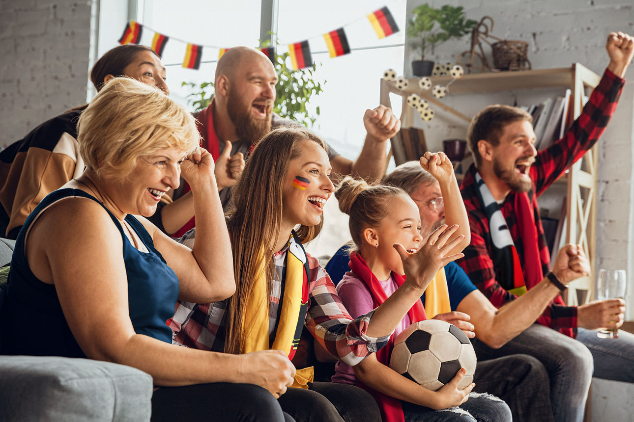 Menschen verschiedener Generationen sitzen in Fußball-Fankleidung auf dem Sofa und jubeln