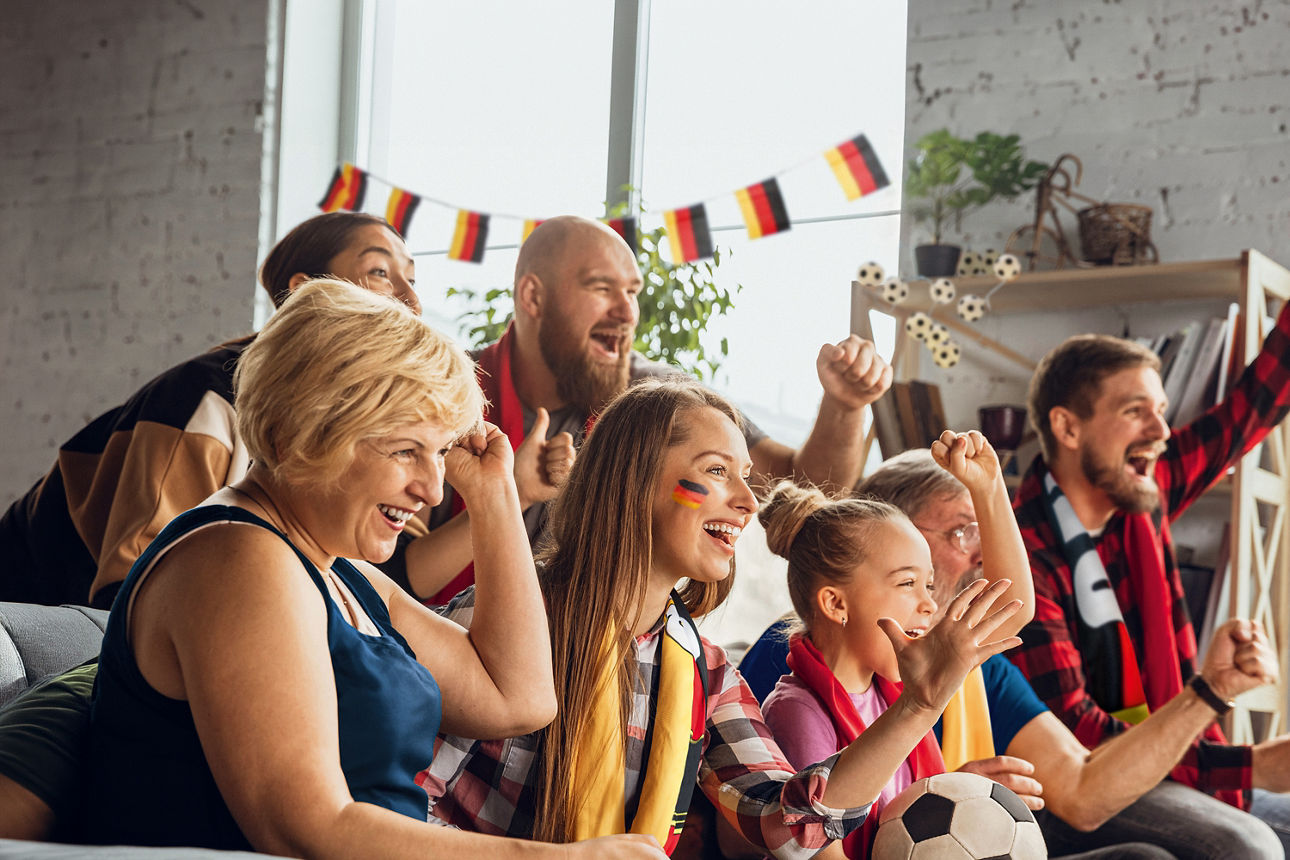 Menschen verschiedener Generationen sitzen in Fußball-Fankleidung auf dem Sofa und jubeln