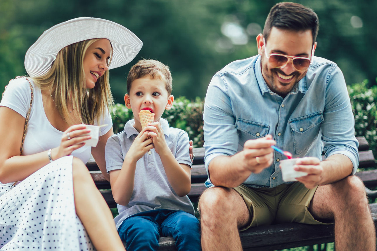 Eine Frau ein Kind und ein Mann sitzen lachend auf einer Parkbank und genießen ihr Eis