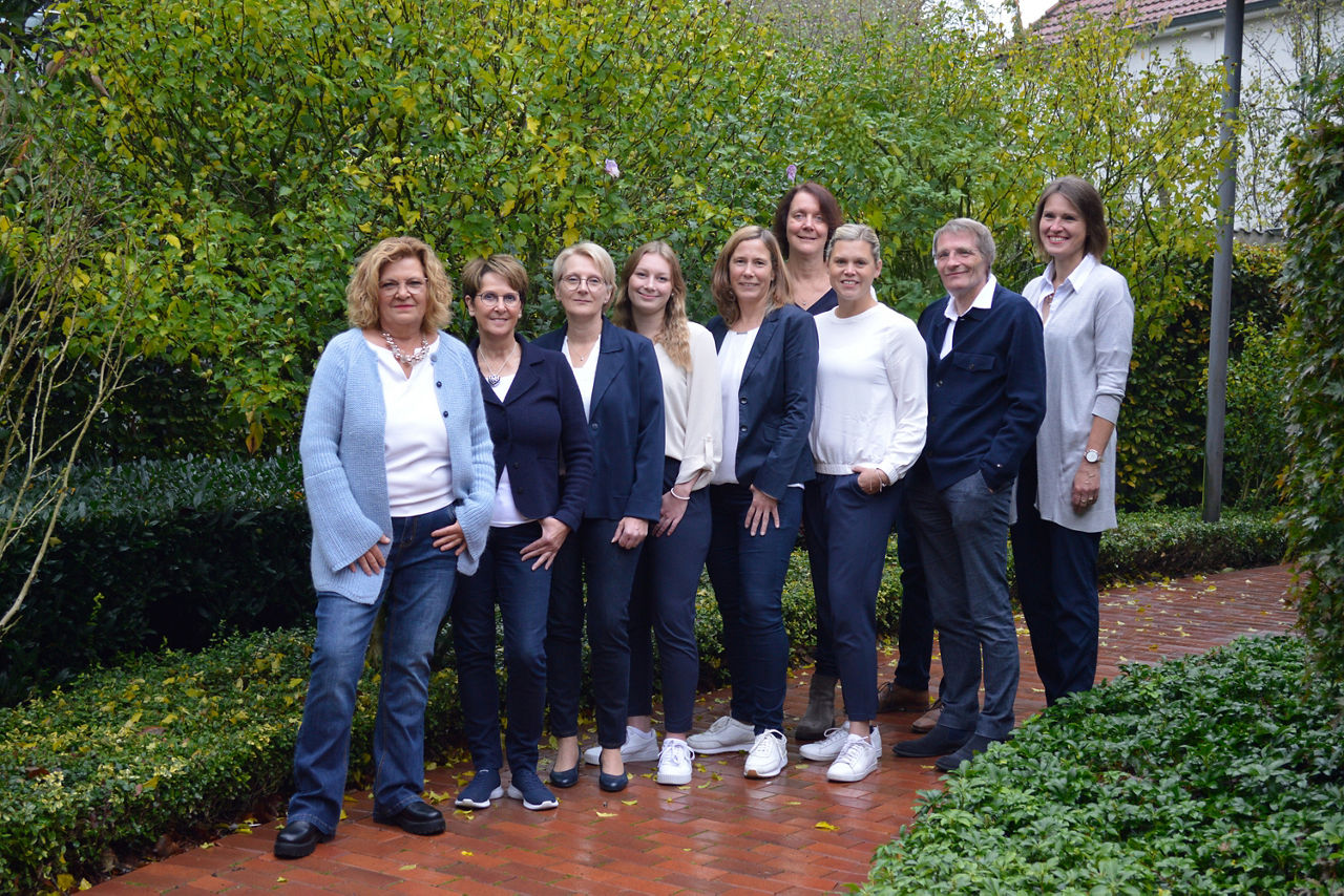 Eine Gruppe von neun Personen steht vor einer üppigen grünen Hecke.