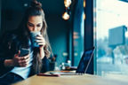 Junge Frau sitzt mit einem Laptop in einem Café und schaut auf ihr Handy