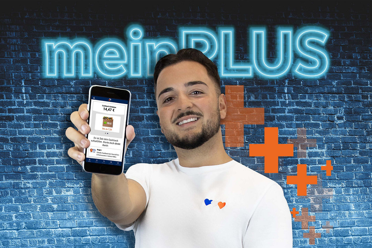 Junger Mann hält Smartphone mit geöffneter App hoch, im Hintergrund leuchtender Schriftzug ‚meinPLUS‘ auf blauer Ziegelwand.