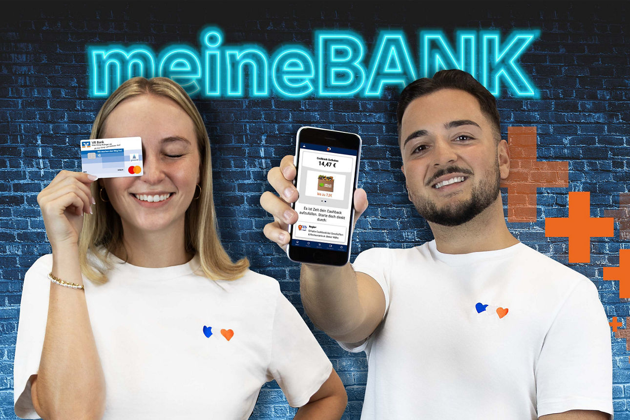 Junge Frau hält Bankkarte vor das Gesicht. Junger Mann hält Smartphone mit geöffneter App hoch, im Hintergrund leuchtender Schriftzug ‚meineBANK‘ auf blauer Ziegelwand.