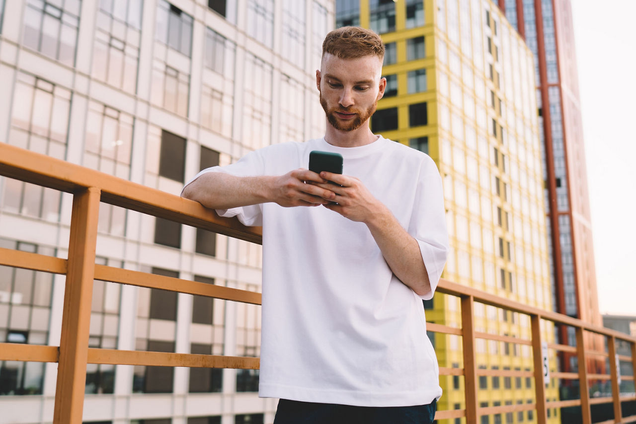 Junger Mann lehnt an einem Geländer und hat ein Handy in der Hand