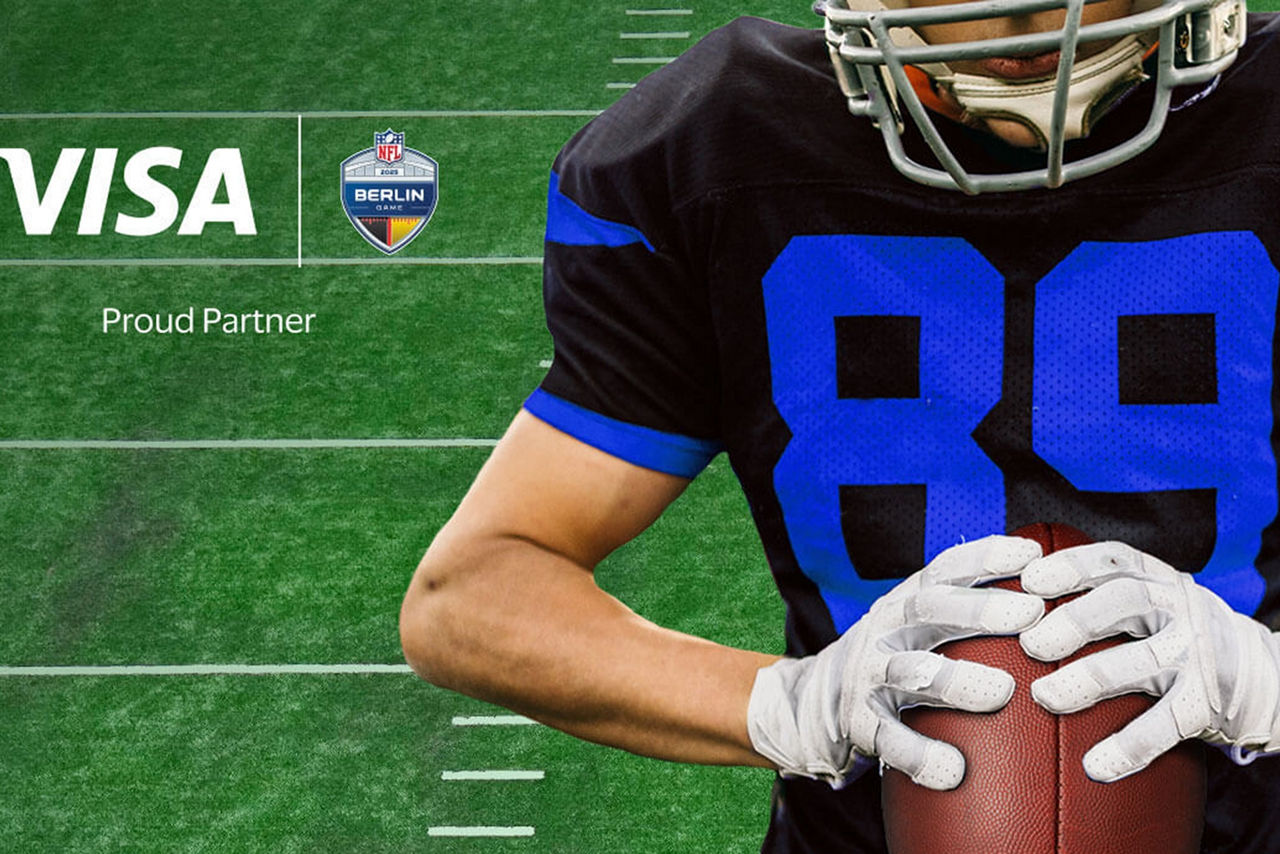 Im Hintergrund ist die Rasenfläche eines Football-Feldes zu sehen. Im Vordergrund sieht man einen Footballspieler in Trikot und Schutzausrüstung mit einem Football in den Händen. Das Gesicht ist nur angeschnitten zu sehen. Im Bild befinden sich folgende Texte: Oben links "VISA Proud Partner NFL" und unten "Touchdown in Berlin". 