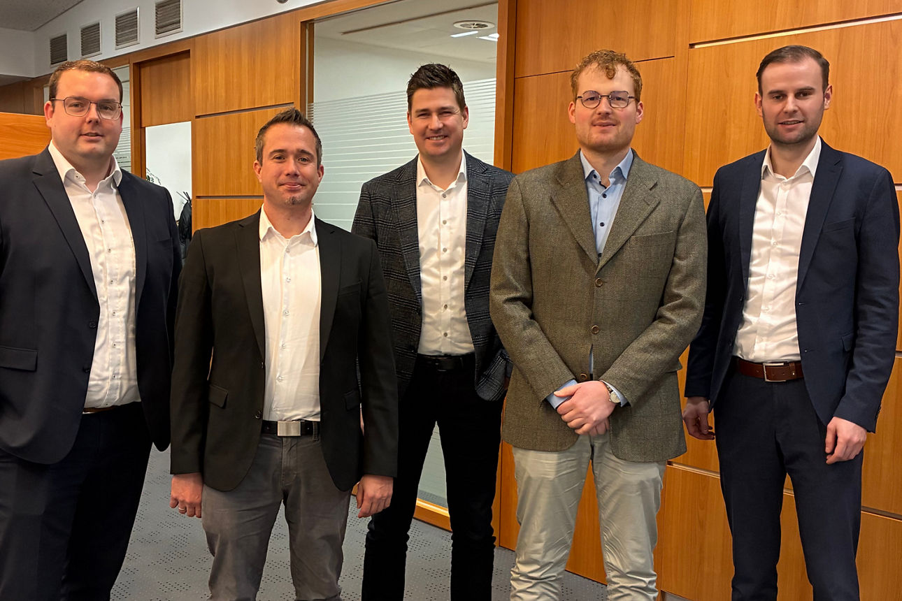 Fünf Männer in Anzügen stehen nebeneinander in einem Innenraum, Gruppenfoto einer Übergabe