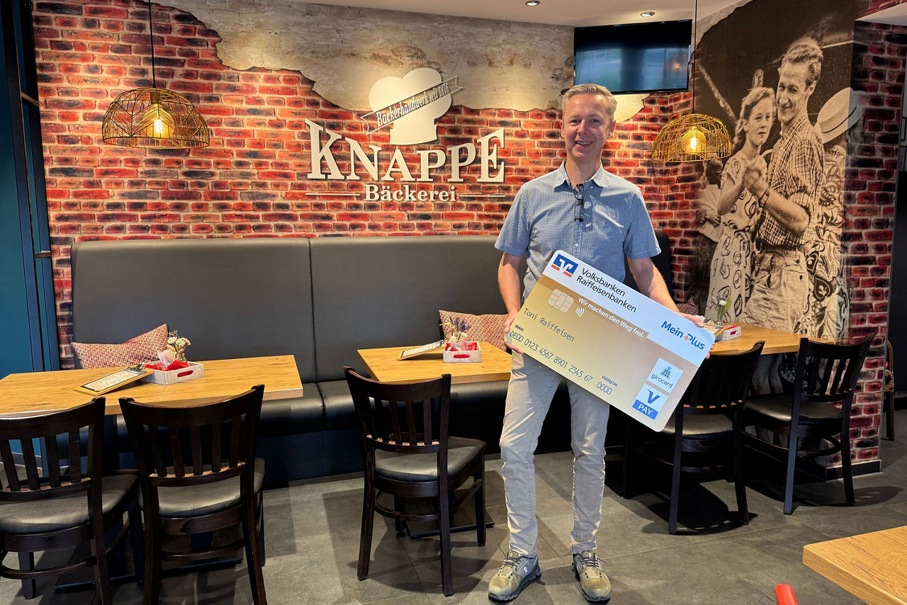 Ein Mann steht in einer Bäckerei vor einer Backsteinwand und hält eine übergroße Volksbanken-Raiffeisenbank-Karte in den Händ