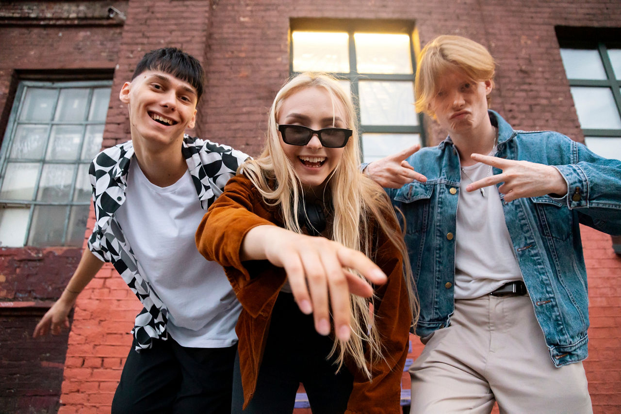 Drei Jugendliche posieren vor einer Backsteinfassade mit großen Fenstern. In der Mitte beugt sich ein Mädchen mit Sonnenbrille zur Kamera, links und rechts stehen zwei Jungen in legerer Freizeitkleidung. Die Aufnahme ist aus einer niedrigen Perspektive fotografiert und vermittelt einen modernen, urbanen Eindruck. Im Hintergrund ist ein Gebäude aus rotem Backstein zu sehen.