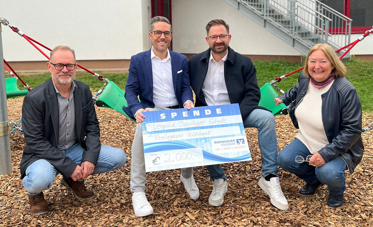 Vier Personen sitzen und knien auf einem Spielplatz mit Hängematten, zwei halten einen großen Spendenscheck über 2000 Euro der Raiffeisenbank.