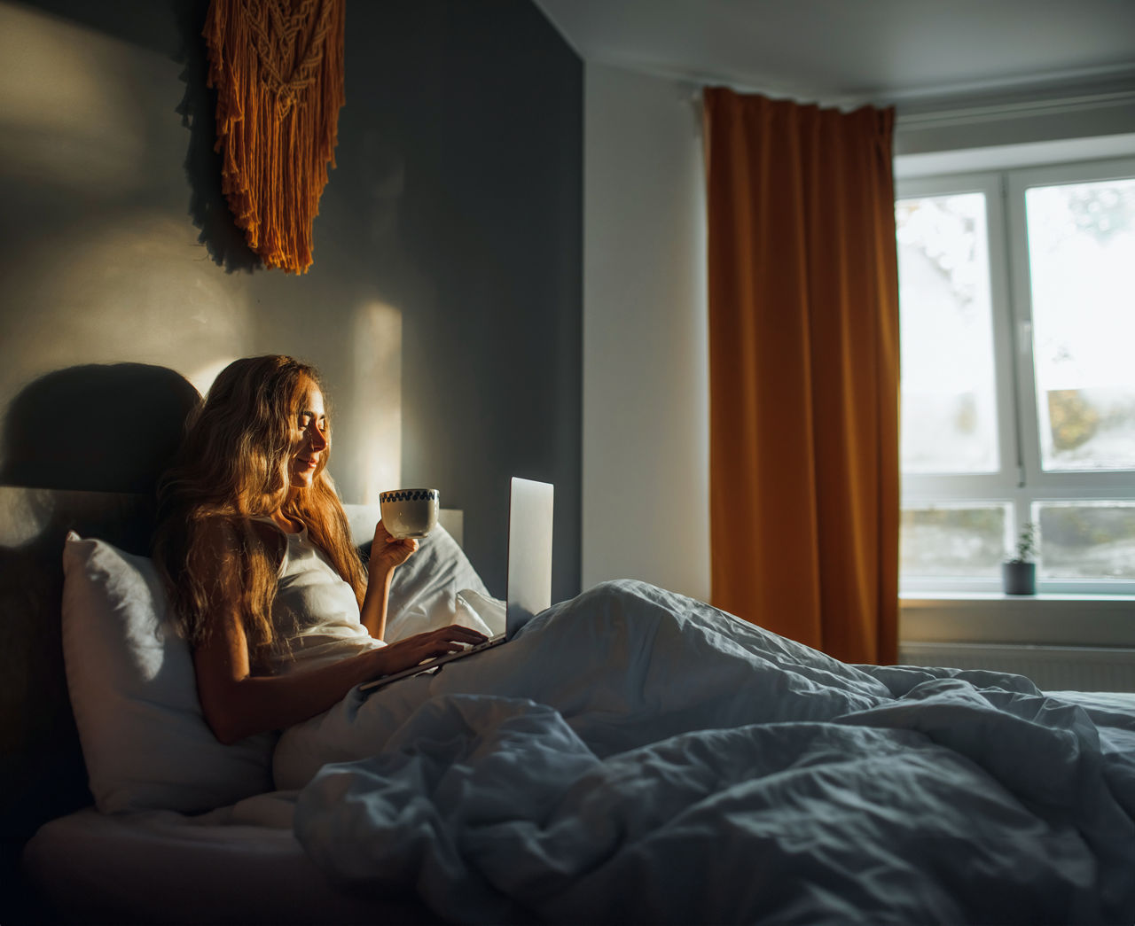 Frau liegt im Bett mit einem Kaffee in der Hand und liest auf dem Laptop Neuigkeiten