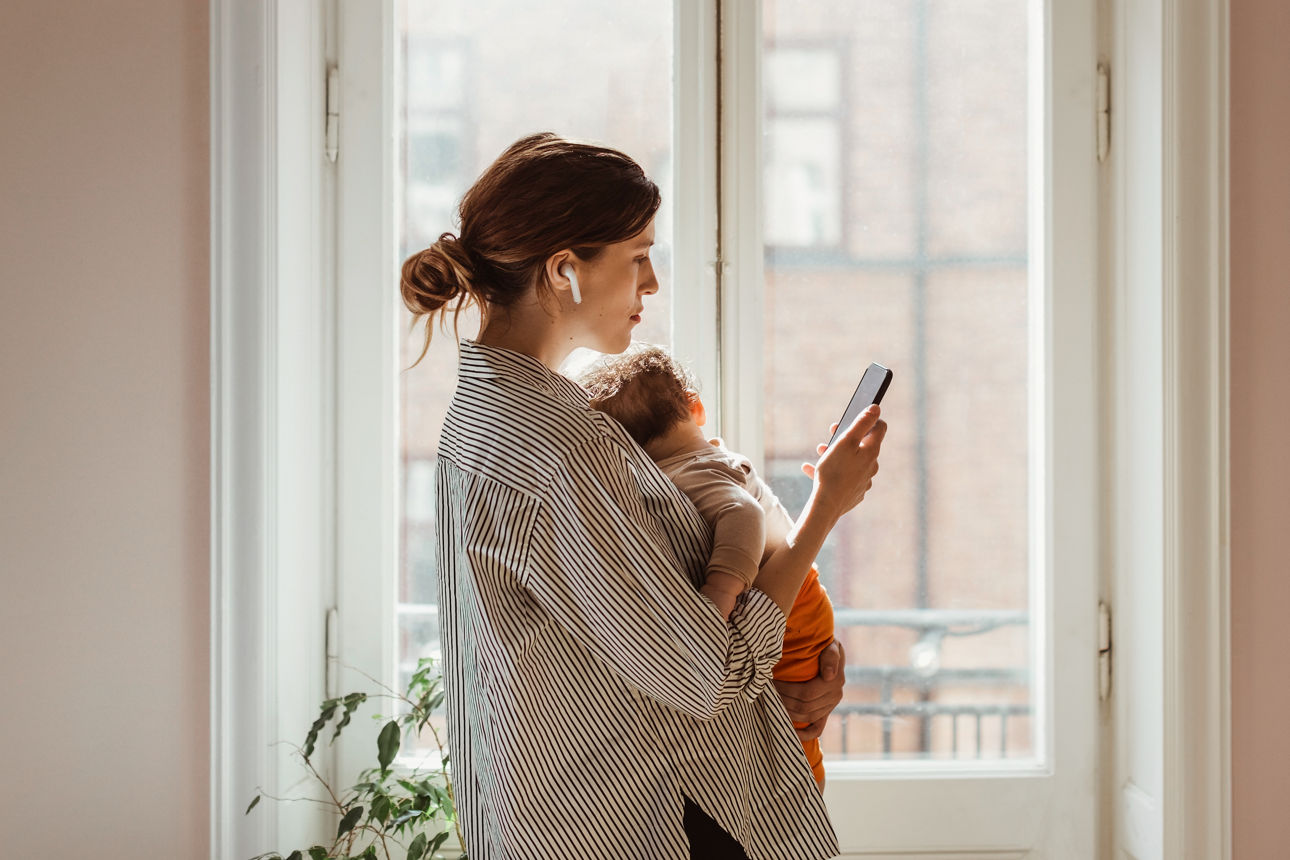 Mama hält ihr Kind auf dem Arm und schaut paraell in ihr Smartphone vor einem lichtdurchflutenden Fenster