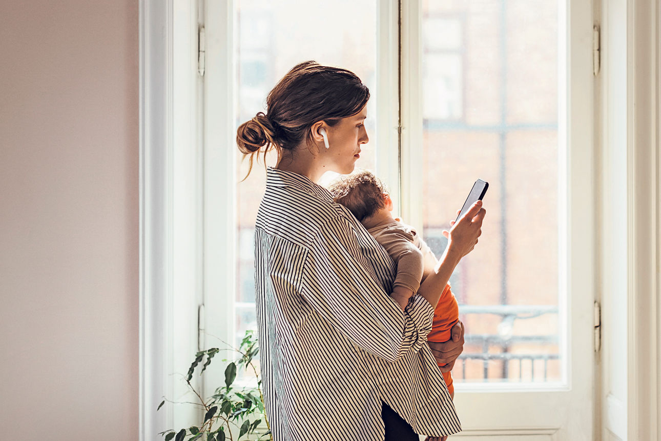 Frau mit Baby auf dem Arm telefoniert via Smartphone und In-Ear-Kopfhörern.
