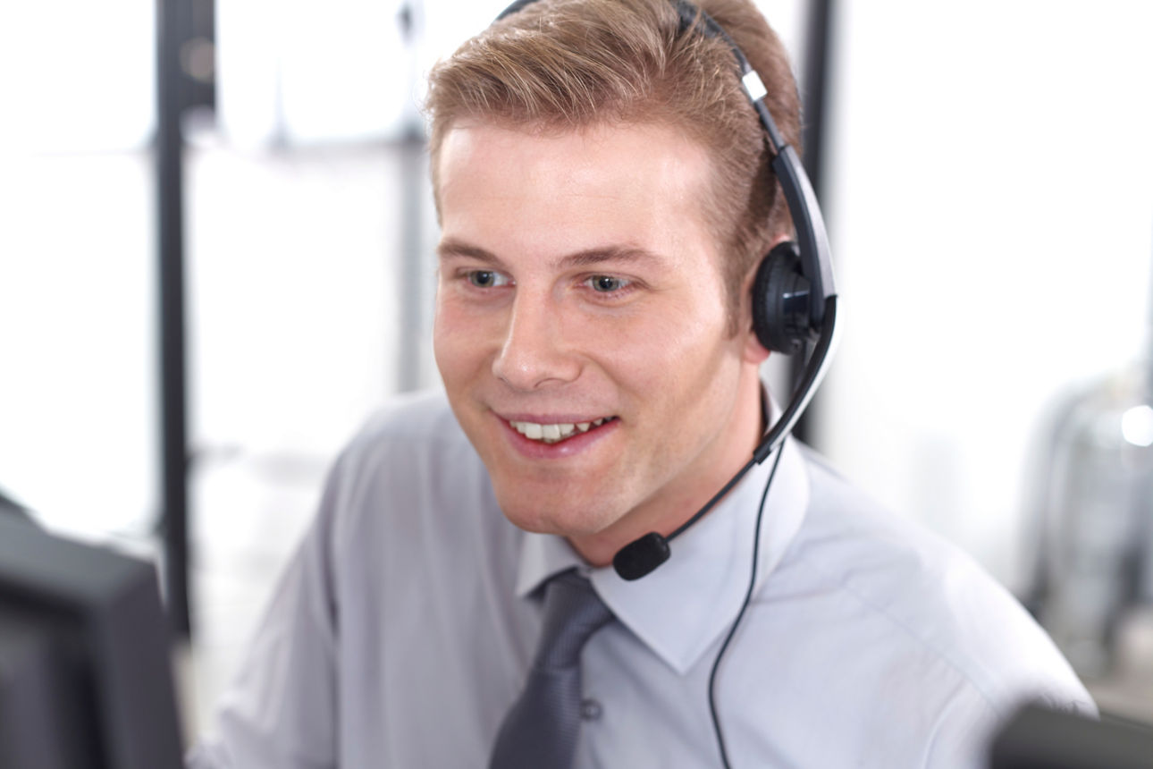 Mann sitzt lächelnd am Schreibtisch und telefoniert mit einem Headset