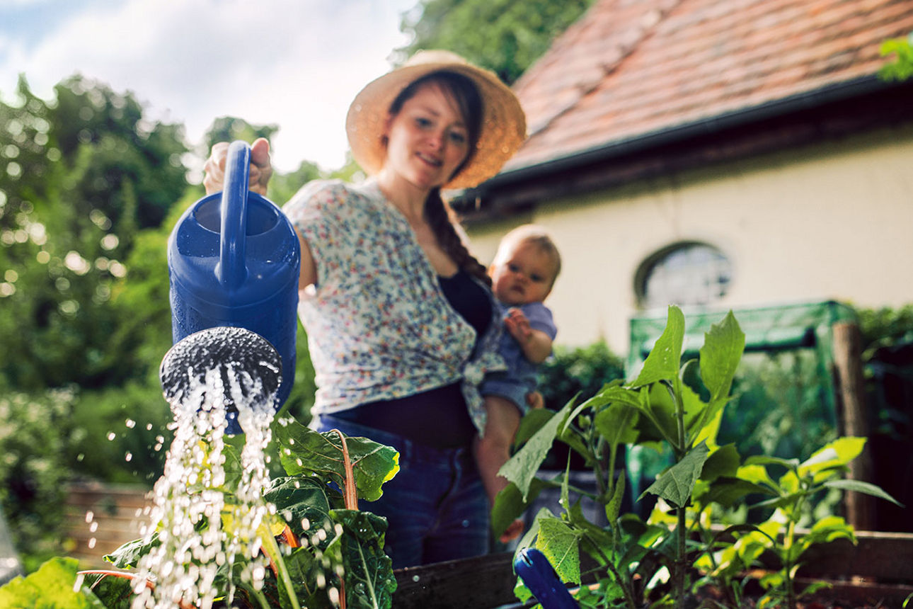 Frau mit Sonnenhut gießt ihre Pflanzen im Garten, während sie ein Baby auf dem Arm hat