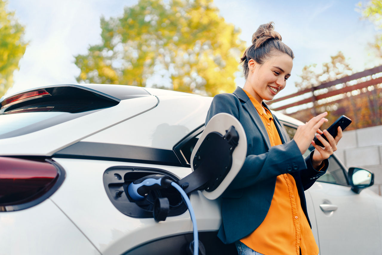 Junge Frau mit Handy in der Hand, lehnt sich an einem Elekto-Auto an. 