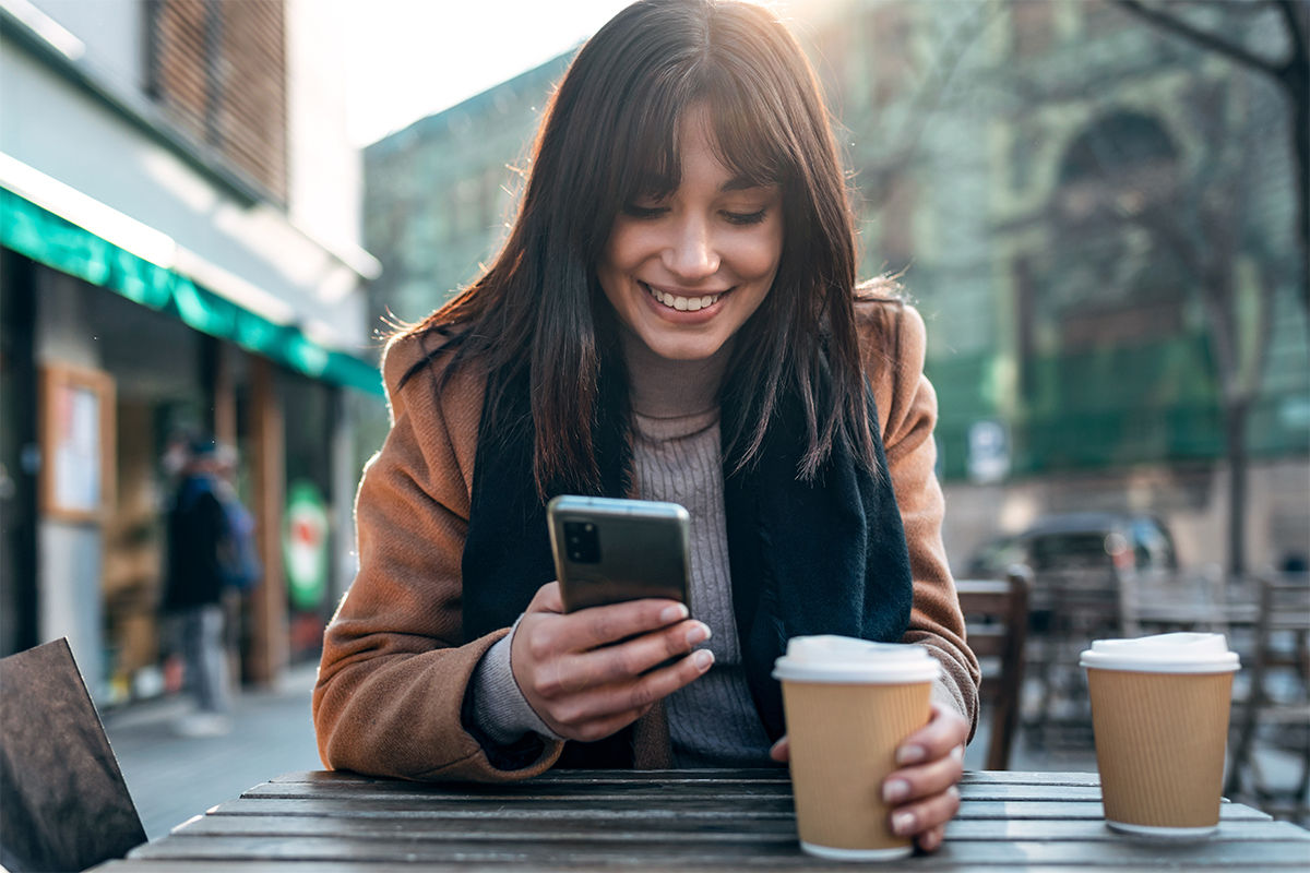 Frau sitzt draußen im Cafe und schaut mit Freude auf ihr Smartphone