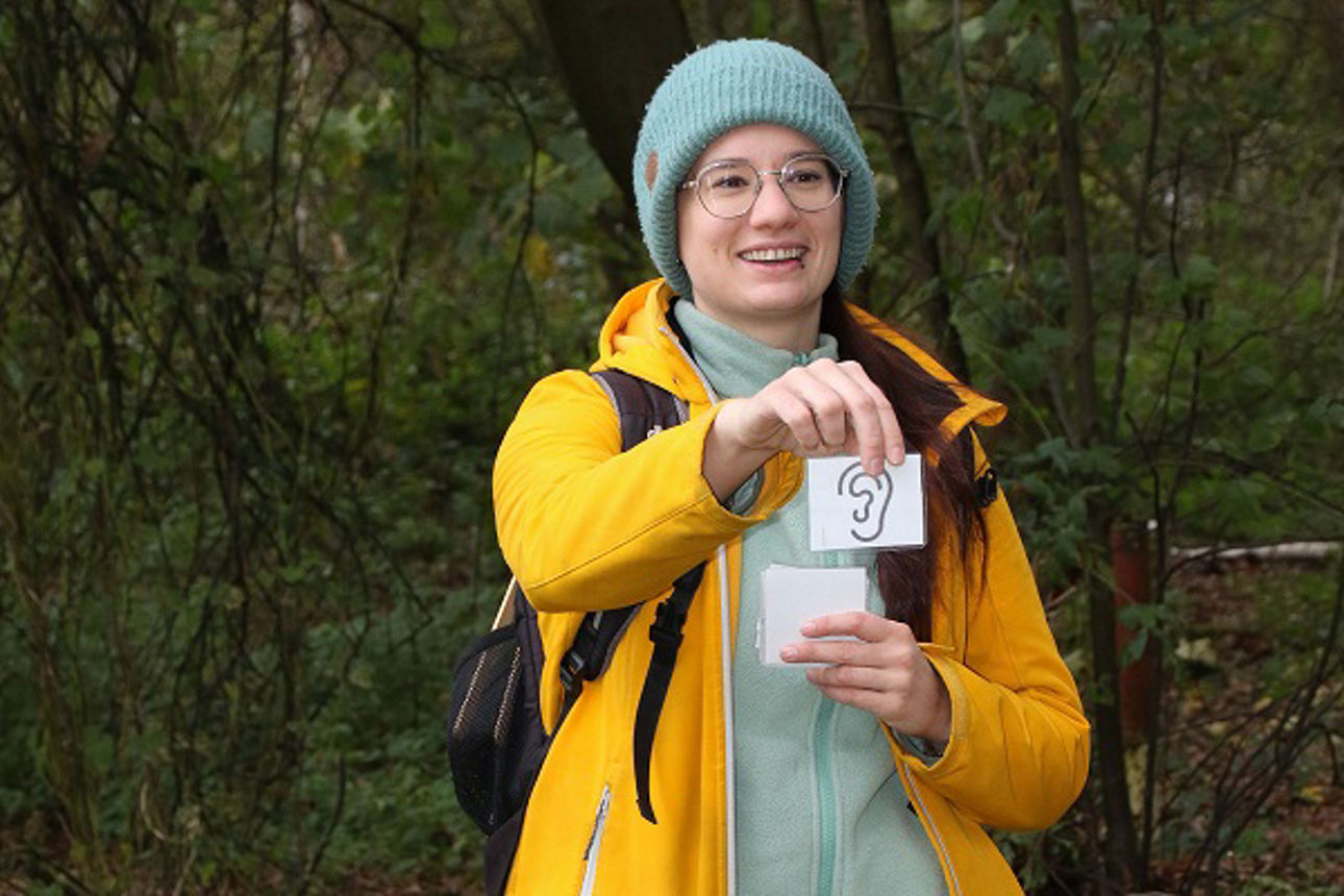 Die Waldpädagogin hält eine kleine Karteikarte, auf der ein Ohr gezeichnet ist, hoch.