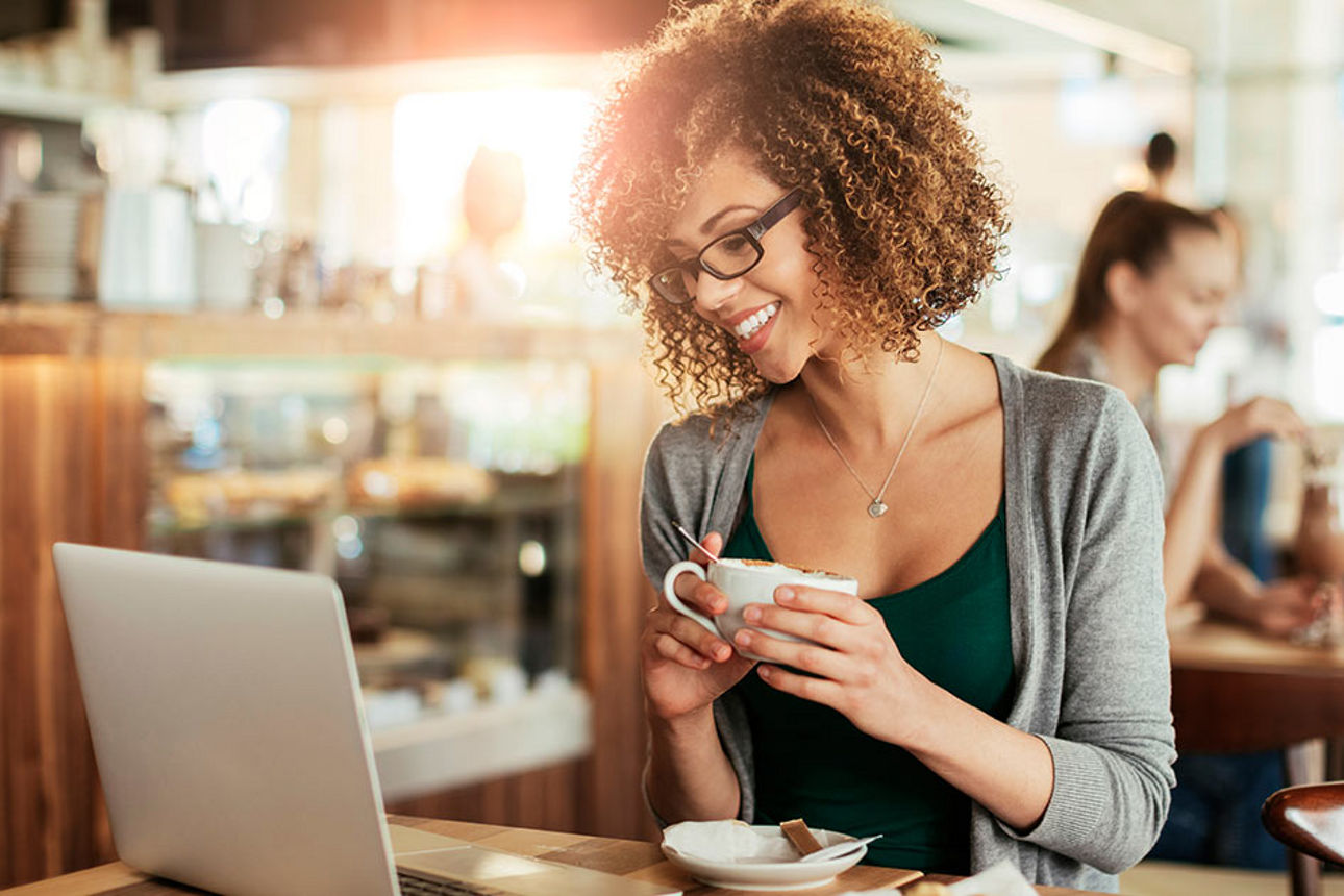Frau mit lockigen Haaren sitzt in einem Café und schaut lächelnd auf ihren Laptop