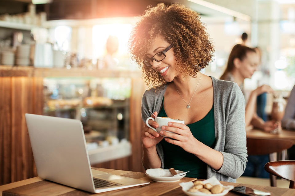 Frau sitzt in einem Cafe und schaut mit einem Kaffee in der Hand aif ihren Laptop