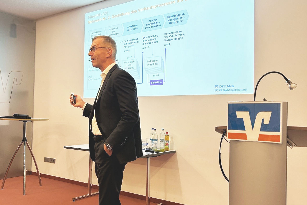 Im Vordergrund ein Redner im Anzug, rechts daneben ein Rednerpult mit VR-Markenzeichen; dahinter an Wand eine gebeamte Seite einer Präsentation der DZ BANK