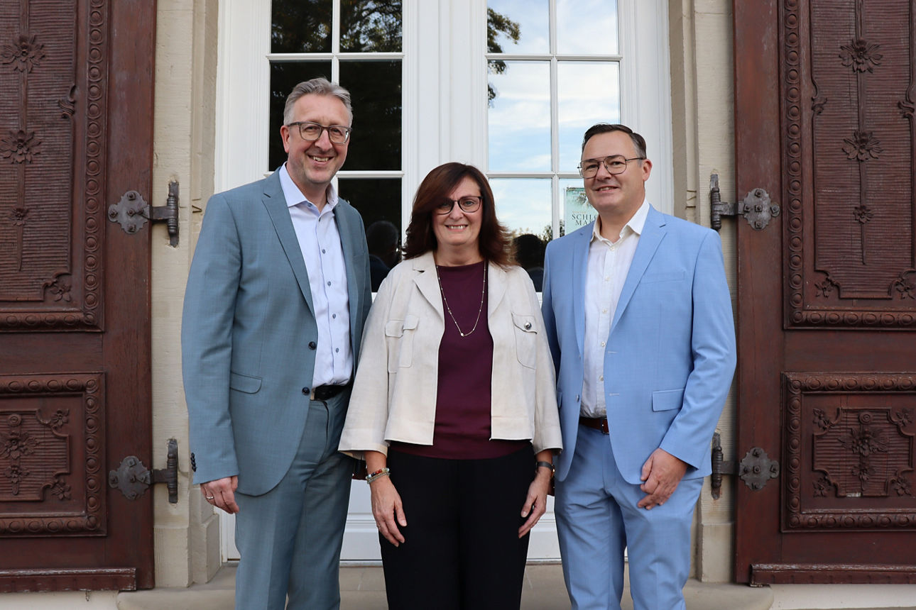 Auf dem Foto ist eine Frau in der Mitte und zwei Männer im blauen Anzug neben ihr. Sie lachen alle in die Kamera