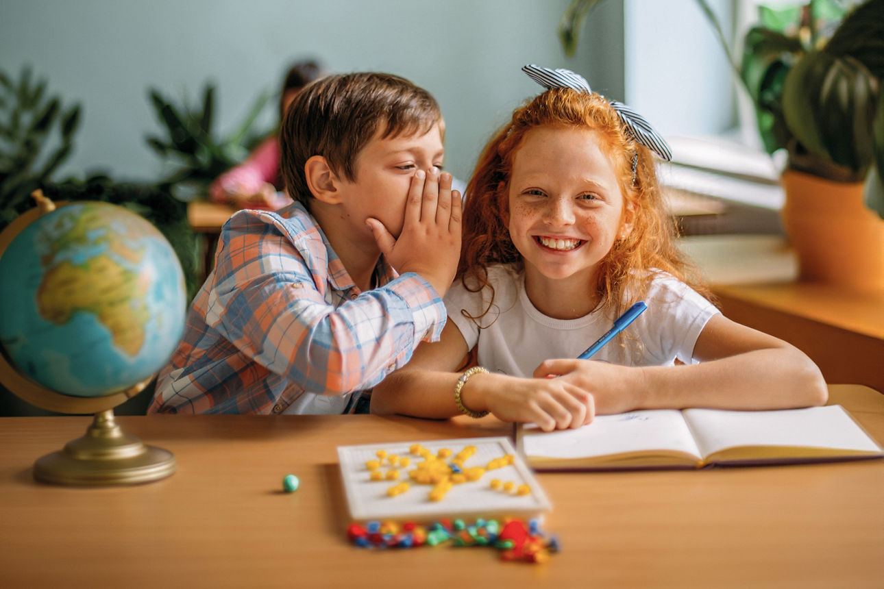 Zwei Kinder sitzen an einem Holztisch mit Schulsachen. Der Junge flüstert dem Mädchen etwas ins Ohr.