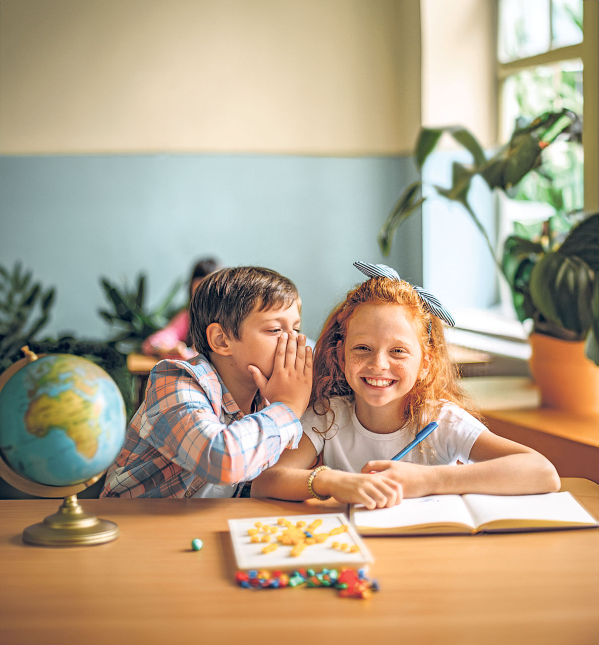 Schüler flüstern sich in einem Klassenzimmer gegenseitig etwas zu. Auf dem Tisch steht ein Globus.