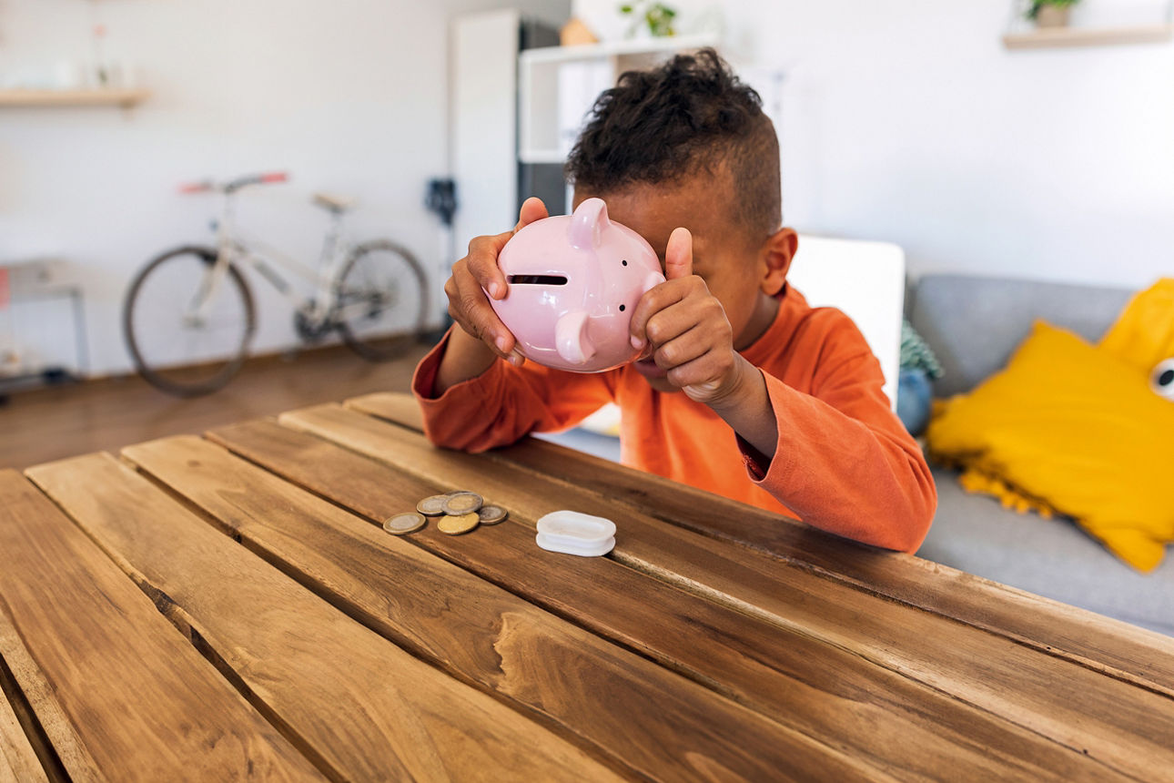 Ein Junge im orangen Pullover schaut am Holztisch in ein rosa Sparschwein, davor liegen ein paar Münzen.