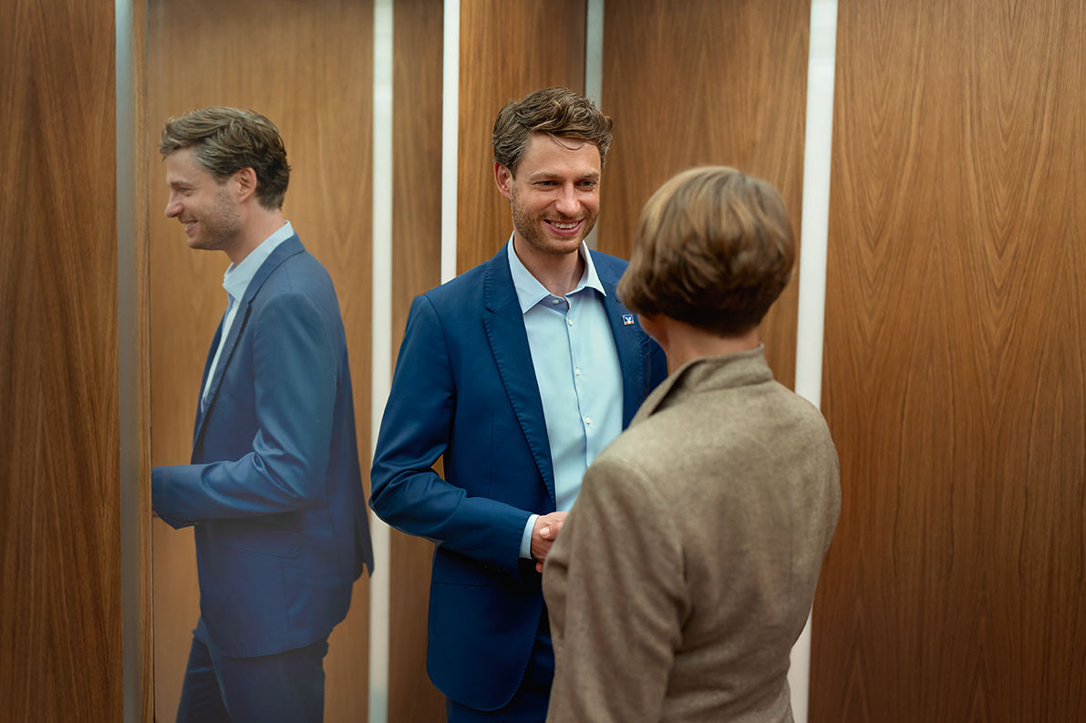 Ein Mann und eine Frau stehen sich lächelnd gegenüber, der Mann spiegelt sich links im Bild